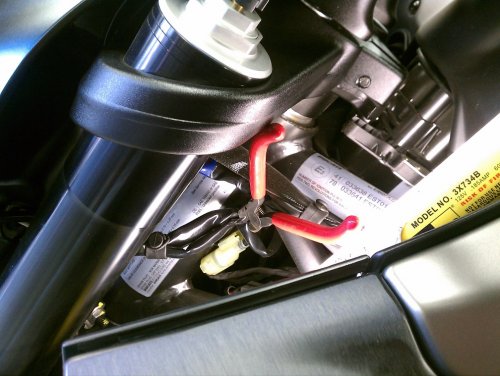 First, remove the two acorn nuts from the bottom of the headlight fairing.  The order is nut, washer, fairing mount, then star-washer.  Strada owners should remove the windscreen for easier access to everything.  Next, snip the zip-tie on the left side that holds the two cables that feed the headlight.  The cable holder is on the frame head.  The two headlight cables are below it and towards the center rear of the frame head.  That yellowish connector you see below the snips is one of them.
